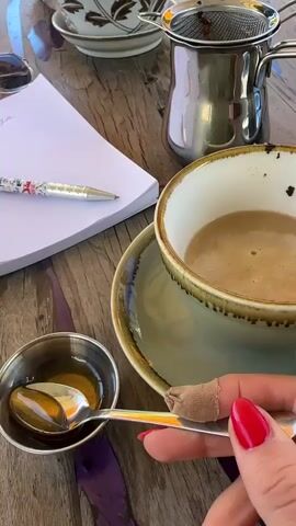 The video shows a close-up of a hand with bright red nail polish holding a spoon. The spoon contains a golden, viscous liquid, presumably honey. The spoon is being brought over a cup of coffee with foam on top. In the background, there is a small metal pot with a strainer, a notepad, and a pen, all resting on a wooden table. The scene appears to be in a cafe or outdoor seating area, suggested by the wooden table and the presence of coffee-related items. The hand is gently tilting the spoon, allowing the liquid to drip into the coffee cup.