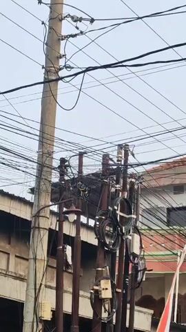 A power transformer is mounted on multiple utility poles. Overhead wires are visible above and around the transformer. Several smaller boxes are attached to the poles below the transformer. Buildings are visible in the background, and the sky is overcast.