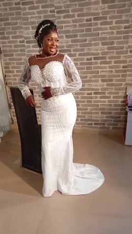 A woman in a white, beaded dress stands indoors, smiling and posing. She is standing in front of a brick wall and leaning against a black chair. She is wearing a beaded necklace and a beaded hairpiece. The room has tiled floors. A curtain can be seen in the background.