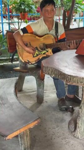 A person is seen from the chest down, playing an acoustic guitar while seated at a rustic table. The individual is wearing a light-colored, striped shirt and blue jeans, with dark brown slip-on shoes. Their left hand is on the guitar fretboard, and their right hand is strumming. The acoustic guitar is made of light-colored wood with dark decorative accents around the soundhole and lower bout.

The camera then pans down to show the person's feet. On the concrete ground between their feet, a small, dark-colored percussion instrument, resembling a foot tambourine or shaker, is visible. The person is tapping this instrument with their right foot, adding a rhythmic element to the guitar playing.

The setting appears to be outdoors or in an open-air covered area, with a textured concrete floor. The table and chairs are designed to look like tree trunks and wood, suggesting a natural or rustic aesthetic. In the background, hints of green foliage and potted plants are visible, characteristic of a garden or cafe environment in Thới Sơn, Vietnam. There is no visible text.