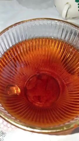 This is a close-up, overhead shot of a clear glass bowl filled with a dark amber liquid, possibly tea or a similar beverage. The bowl has a ribbed texture and a gold rim. The liquid is translucent, allowing light to pass through and highlight the color. There is a noticeable reflection or glint of light on the surface of the liquid. The bowl sits on a white, possibly tablecloth, with a hint of a patterned edge visible at the bottom. In the upper right corner of the frame, a portion of a white mug with a handle is visible, suggesting the beverage might be prepared for drinking. The lighting appears to be neutral, and there are no distinct cues for time of day or weather. The overall impression is one of simple domesticity, perhaps a moment of quiet preparation or enjoyment of a warm drink. The setting seems to be indoors, in a home environment. There is no visible text in the media.