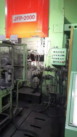 The video shows a large industrial machine with a red panel labeled "JFP-2000" at the top. The machine is green and has a control panel with several buttons and switches on the right side. There are various wires and hoses connected to the machine, and a metal cage on the left side. The machine appears to be operating, with sparks or flashes of light visible in the central area. The environment is a factory floor, with a dark, textured floor. The video is filmed from a medium shot, facing the machine.