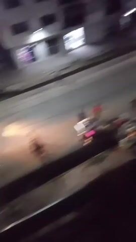 The video shows an outdoor scene at night, viewed from an elevated position, likely a balcony. The foreground shows a section of a balcony railing, obscuring part of the view.

Below, on a street, a group of people are gathered. Some appear to be standing near a pile of items, possibly debris or belongings. Others are walking or running across the street.

Vehicles are visible on the street, including what seem to be auto-rickshaws or similar small motorized vehicles, with their headlights on. The background shows a building with illuminated windows and minimal details are visible. The street is wet, reflecting some of the light.

The overall lighting is dim, with artificial light sources creating some illumination. The video appears to be taken with a mobile device, and some camera shake is evident.