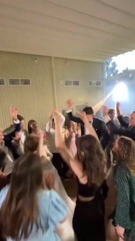 A group of young people are dancing and celebrating in a dimly lit room. The room appears to be a hall or event space with a high ceiling and white walls. There are lights set up, creating a festive atmosphere. Some of the individuals are wearing formal attire, like tuxedos and dresses, suggesting a special occasion such as a prom or wedding reception. The camera is positioned to capture the energy of the dancing crowd, with many arms raised in the air. There are also some hints of technology in the background, including what appears to be a camera on a tripod and possibly a screen. The overall scene depicts a lively and joyous gathering.