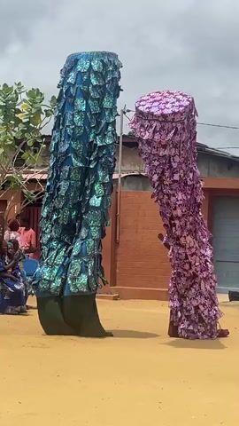 Two figures, dressed in tall, conical costumes covered in layered, reflective fabric pieces, stand facing each other on a dirt ground. The figure on the left is predominantly blue, while the figure on the right is predominantly purple. Their heads are obscured by the costumes, which are tall and tapered, reaching from the ground to above the visible heads. The fabric pieces appear to be square or rectangular, with a patterned design. The figure on the right has dark, visible hands extending from the costume. To the left of the figures, a group of people are seated, some of whom are watching the figures. Behind the figures, there are buildings with red brick and corrugated metal roofs. The sky is overcast.