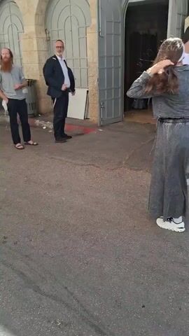 The video opens with a woman in a long, dark denim dress and white sneakers standing with her back to the camera, her hair tied up with a white bandana. She is facing away from a large, arched doorway and several people are walking past her. To her right, a man in a white t-shirt, dark pants, and black sneakers walks away from the camera. Behind him, another young man in a white t-shirt and khaki shorts holds a water bottle, and a third young man in a red t-shirt and black shorts walks with him. An older man in a dark suit and glasses stands near the doorway on the left. The scene is outdoors, with a paved ground and a building with stone walls and arched entrances. The camera then pans to the right, showing a young man in a white t-shirt and khaki shorts holding a water bottle, followed by another young man in a red t-shirt. Further down the street, a woman in a pink dress walks by a trash can, while a young man in a white t-shirt and a white cap stands near a wall, reading a book. Another person in a long black coat and hat stands near the trash can, also looking at papers. The camera then moves further down a street with arched stone buildings and an overhead canopy. A group of young men, mostly wearing t-shirts and shorts, are walking away from the camera. Some are wearing hats, including one with a kippah. The video ends with the group continuing down the street.