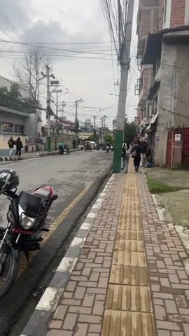 The video shows a street scene with a sidewalk on the right and a road on the left. Several people are walking on the sidewalk and riding motorcycles on the road. The sky is overcast. There are buildings on both sides of the street. A motorcycle is parked on the left side of the road in the foreground.