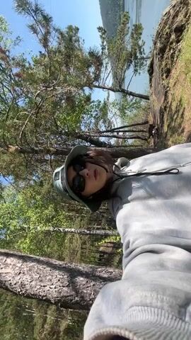 A vertical video shows a young person in an outdoor, natural setting, taking a selfie video. The individual is centered in the frame, wearing dark sunglasses and a light green or grey bucket hat, along with a grey hooded sweatshirt. They have short, dark hair.

Initially, the person looks directly at the camera, then shifts their gaze to the right, looking out over the landscape. The background features a vibrant natural environment. On the left and in the foreground, there are numerous trees, including pines with visible bark and some deciduous trees with bright green leaves.

Behind the trees and to the right of the person, a large body of water, likely a river or lake, is visible under a clear, bright blue sky. The water appears calm, reflecting the light. On the far side of the water, distant hills or mountains can be seen. The ground near the water's edge appears somewhat rocky or grassy. The overall impression is one of a sunny day in a serene natural landscape, likely a forest bordering a large body of water. The location is Sliznevo, Russia. No text is visible in the video.