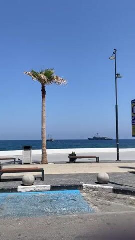 A palm tree stands on a paved walkway overlooking the ocean under a clear blue sky. In the distance, two boats are visible on the water. A modern ship is further out, while a smaller, older-looking sailing vessel is closer to shore. A lamppost with two lights stands to the right of the palm tree. In the foreground, a parking space is marked with blue and white lines. The overall scene appears to be a coastal town or promenade on a sunny day.