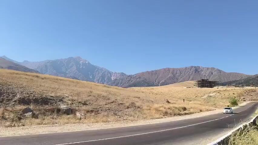 The video begins with a view of a winding road through a dry, hilly landscape under a clear blue sky. A white car is seen driving on the road. In the distance, mountains rise, and a partially constructed building is visible on a hilltop. As the video progresses, the camera pans to the right, revealing wind turbines and a power line tower on a ridge. The landscape continues to be dry and sparsely vegetated. Further on, the camera captures a wide expanse of a large, turquoise body of water, likely a lake or reservoir, nestled between mountains. The opposite shore of the water features a town or village with scattered buildings and greenery. The camera continues to pan, offering a panoramic view of the water and the surrounding mountainous terrain. The scene is bright and sunny, suggesting daytime. The video appears to be a travelogue or scenic recording of a journey through a mountainous region with a prominent body of water.