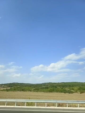 A street lamp stands tall against a clear blue sky with scattered clouds. A metal guardrail runs horizontally across the foreground, separating a road from a rural landscape. The landscape features rolling hills covered in green vegetation and patches of dry, brown earth. The camera appears to be moving, as indicated by the slight blur and shift in perspective. There are no people visible in the video. The overall scene is one of a roadside view on a sunny day.