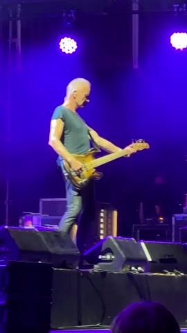 A man with short, gray hair and a dark blue t-shirt and black pants is playing a bass guitar on a stage. He is on the left side of the frame, facing right and looking down at the guitar. He is standing on a platform with several large speakers in front of him. Behind him, there are bright purple lights and some stage equipment. The background is dark.