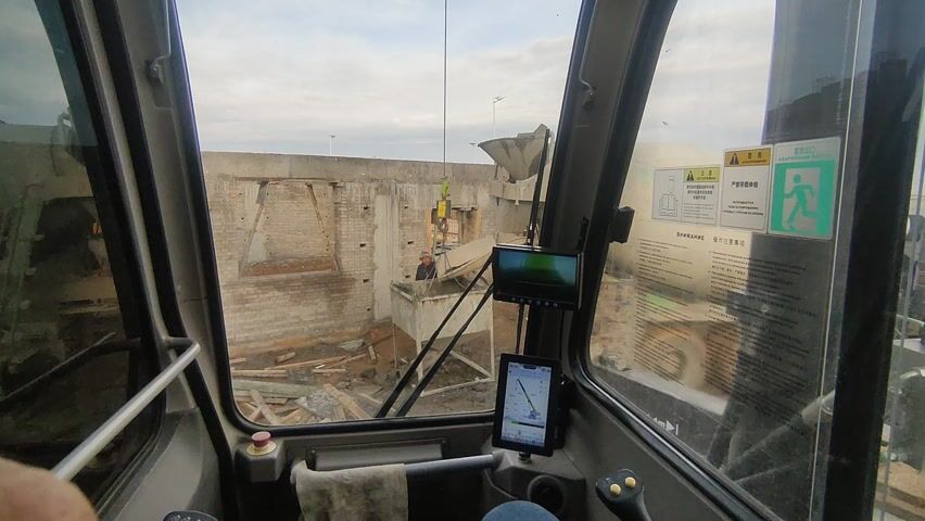 The video shows the view from inside the cab of a crane or other construction vehicle. Through the windshield, a concrete mixer truck is visible, pouring concrete into a large, metal container. A man in a dark jacket and hat stands nearby, overseeing the process. The construction site is littered with debris, including broken bricks and lumber. A monitor and a tablet are mounted on the interior of the cab, displaying information related to the vehicle's operation. The sky is overcast.