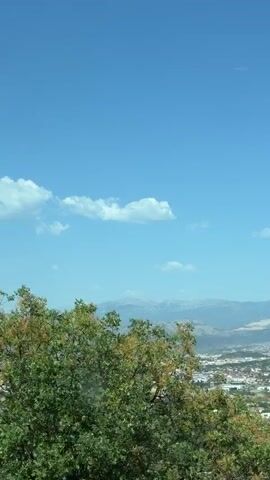 The video displays a scenic overview of a town nestled in a valley, with mountains in the background under a clear blue sky dotted with a few white clouds. The foreground features lush green trees and foliage, some of which partially obscure the view of the town below. The town itself appears to be a mix of residential areas with houses, and some larger buildings or facilities, possibly including a stadium or sports complex with a red roof visible. The overall impression is one of a peaceful, rural or semi-rural setting on a bright, sunny day.

The time of day appears to be daytime, given the bright sunlight casting shadows and illuminating the landscape. The weather is fair and clear, indicated by the blue sky and visible clouds. The location, Liješnje, Montenegro, suggests a Balkan landscape with hilly terrain and significant vegetation. There is no visible text within the video. The scene evokes a sense of tranquility and natural beauty, with the vastness of the sky and mountains contrasting with the organized, yet somewhat spread-out, human settlement. The movement in the video suggests a slow panning motion, likely capturing a panoramic view from a higher vantage point.