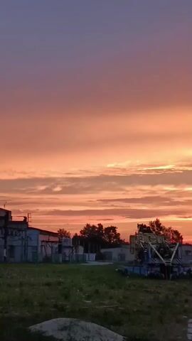 The video shows a sunset with orange and purple clouds over an industrial area. There are several buildings, including a large arched structure, and a tall pole with lights. In the foreground, there is a pile of sand and some vegetation. A piece of machinery with a crane is visible on the left. The road is paved in parts and dirt in others.