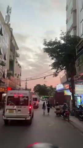 The video shows a street scene with a pink and grey sunset sky. On the left is a multi-story building with air conditioning units. There's a white van with "CAT BA VENTURES" on its side, followed by another van of the same model. People are walking along the sidewalk. A scooter with a large load is on the right. Power lines run overhead.