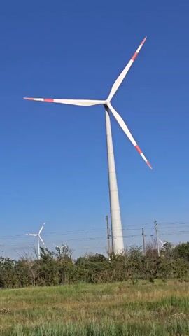 A video shows a wind turbine against a clear blue sky. The turbine has three white blades with red tips. In the background, other wind turbines are visible, along with trees and power lines. The foreground is a field of green grass and some dry brush. The camera is moving slightly, capturing the slow rotation of the turbine blades. The overall impression is one of a rural landscape with renewable energy infrastructure.