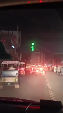 The video, shot from the perspective of someone in a vehicle, shows nighttime street traffic. The scene is dimly lit by streetlights and vehicle lights. Several auto-rickshaws and a large truck are visible in the foreground. The camera focuses on the back of the truck, and behind it, a mosque minaret is illuminated in green. People can be seen walking along the side of the road. The vehicle the camera is in is moving slowly through traffic. The street is paved and lined with buildings.