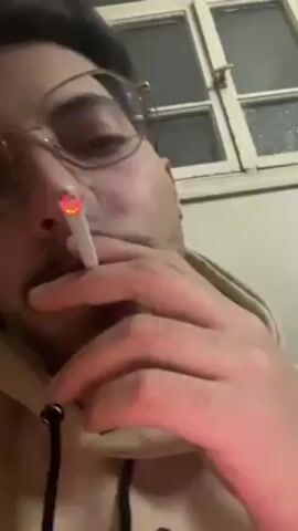 A man wearing glasses and a beige hoodie is smoking a cigarette. He is looking directly at the camera. There is a window in the background. He exhales smoke.