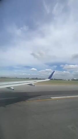 The video shows a view from inside an airplane on a runway. The wing of the plane is visible with its blue-tipped tail. The runway is gray with yellow lines and dark tire marks. Beyond the runway is a grassy area and buildings in the distance under a blue sky with scattered white clouds. The plane appears to be stationary.