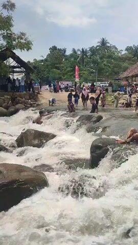The video shows a lively scene at a river or waterfall, with many people enjoying the water. In the foreground, several children are playing in the fast-flowing water, sitting on rocks and being splashed by the white foam. One boy in particular is prominently featured, sitting on a large rock in the middle of the rapids, looking towards the camera. Behind the children, a crowd of adults and children are gathered on the riverbank, some standing and observing, while others appear to be preparing to enter the water or have just come out. There are several structures in the background, including a rustic-looking building with a thatched roof and another partially visible building with a similar roof. Trees and lush greenery surround the area, suggesting a natural, outdoor setting. The sky is partly cloudy. Some people are wearing colorful clothing, and there are signs visible in the distance, indicating this is likely a recreational area or tourist spot. The overall atmosphere appears to be one of fun and enjoyment.