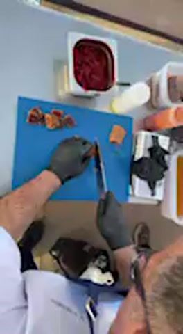 The video provides an overhead view of a person preparing food on a bright blue cutting board. The individual, wearing a white short-sleeved shirt and black gloves, is seen from the chest down, expertly slicing pieces of what appears to be dark red meat or fish, likely tuna, given the Barbate context. Several pre-cut rectangular portions are already arranged on the board, and the person uses a sharp knife to make precise, skilled cuts, further dividing the food into smaller, uniform pieces.

The scene suggests a professional food preparation area, possibly a food stall or an outdoor culinary event. In the background, a stainless steel counter holds various containers: a square container with a dark red substance (possibly a sauce or dip), a white squeeze bottle, and several white plastic containers, some holding orange or brownish contents. The setup appears organized and clean.

Spanish dialogue is audible in the background, with one voice asking, "Estáis cortando más porque lo vais a dar a probar?" (Are you cutting more because you're going to give it to try?). This indicates that the prepared food is intended for tasting or distribution, suggesting a public event or demonstration. The bright lighting illuminates the food preparation process clearly. There is no visible text in the video frame. The overall activity conveys a sense of culinary skill and a lively atmosphere.