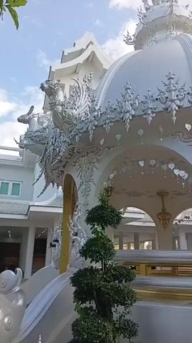 The video shows a white domed structure adorned with intricate carvings and sculptures. The structure is situated outdoors with a cloudy sky visible in the background. A green bush is positioned in the foreground. The camera pans slowly, revealing more of the architectural details of the white structure, including what appear to be animal figures and decorative motifs along the roofline and supporting arches. Several other white buildings with similar architectural styles are visible in the distance.