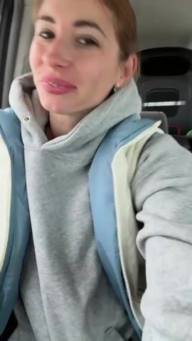 A woman is sitting in the passenger seat of a car. She has brown hair pulled back in a ponytail, fair skin with a rosy complexion, and is wearing a gray hooded sweatshirt with a light blue and white vest. She is smiling and talking. She is wearing braces. Behind her is the back of the front seat, and glimpses of an airport can be seen through the window. In the background a child is talking and sounds of a vehicle are audible.