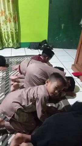 A group of young boys, dressed in traditional Islamic attire, are on their knees on a patterned rug. Some are bowing with their foreheads touching the floor, a posture commonly used in Islamic prayer. One boy in the foreground is looking towards the camera and has his hand on his head. Another boy behind him is also looking forward. The room has light green walls and is dimly lit. In the background, there are curtains and some shelves. The scene appears to be indoors, likely in a place of worship or a dormitory setting.