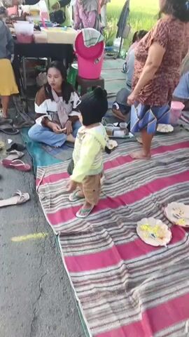 A group of people are gathered at an outdoor market. A young child in a yellow and white striped jacket and brown pants is standing on a striped blanket. The child is holding a black plastic bag and pointing to the left. A woman in a brown and white top and blue jeans is sitting on the ground and looking at the child. Several other people are standing and walking around in the background. There are tables with various items on display, including food and drinks. The ground is covered with a blanket and several pairs of sandals. There are also some yellow lines on the asphalt. The environment appears to be a street or roadside market.