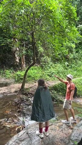 Two individuals, a woman and a man, are standing by a stream in a lush, green forest. The woman is in the foreground, facing away from the camera, wearing a dark green poncho and red socks. She is leaning slightly forward, looking towards the stream. The man is to her right, facing her, wearing a light-colored hat, an orange t-shirt, and beige shorts. He is also looking towards the stream and appears to be gesturing with his right hand. The stream flows over rocks, and the surrounding area is covered in dense vegetation, including trees and bushes. The ground appears to be damp, suggesting it may have recently rained.