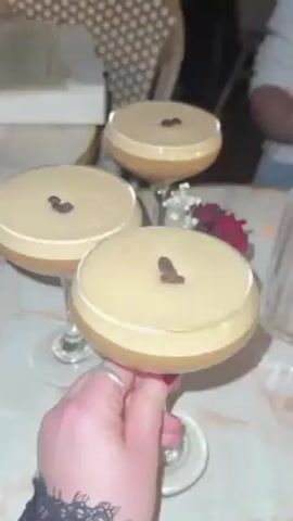 The video shows three coffee martinis in stemmed glasses, each topped with three coffee beans. The drinks sit on a marble-patterned table in what appears to be a restaurant or bar. A hand with a silver ring is holding one of the martini glasses. Background details include a chair, other people, and a small floral arrangement.