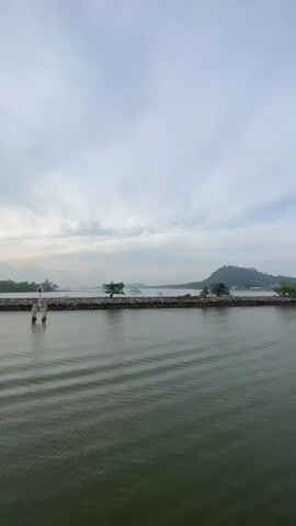 The video shows a calm sea under a partly cloudy sky. A long stone breakwater stretches across the water with a small structure on it. There are trees on the breakwater and forested islands in the background. The sun is breaking through the clouds, creating a reflection on the water. The camera is on a boat that is moving away from the breakwater. A person is briefly seen holding a phone to take a picture. A small boat passes in the distance.
