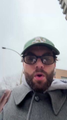 A man is recording a selfie video outside. He is wearing glasses, a green baseball cap with the Oakland A's logo, and a gray coat over a hooded sweatshirt. He has a beard. A tall building is visible in the background. The sky is overcast. It appears to be snowing lightly. The man is walking and talking, and his mouth is open.