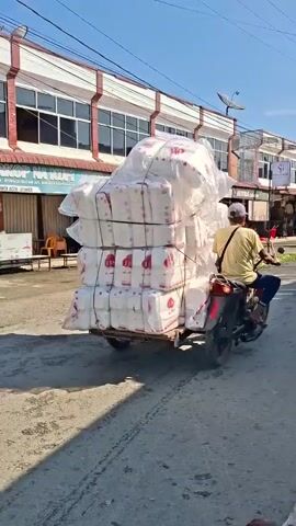 A man wearing a yellow shirt and a hat rides a scooter with a trailer. The trailer is loaded with many large white bags, some stacked in boxes. The scooter is on a paved road in front of a row of buildings with storefronts. Another man, wearing a white shirt and a hat, is seated on the side of the road, looking away from the camera. Another man on a motorcycle passes by. The sky is blue.