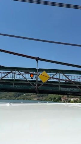 The video shows a perspective looking up at a bridge from below, possibly from a boat or vehicle traveling underneath it. The bridge is made of green painted metal girders and supports, with some signs of age and wear, including peeling paint and rust. A bright orange traffic light is visible on the side of the bridge, indicating a stop signal. A yellow diamond-shaped sign is also mounted on the bridge structure, and a small white security camera is attached to one of the supports. The bridge spans over a body of water, likely a river, with lush green hillsides dotted with buildings and vineyards in the background, suggesting a rural or semi-rural European setting, consistent with Reventin-Vaugris, France. The sky above is a clear, bright blue, indicating a sunny day. The overall impression is of infrastructure in a scenic, natural environment. There is no visible text in the media file.