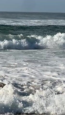 A video captures a close-up view of ocean waves breaking on a beach. The camera is positioned low, near the water's edge, and focuses on the churning foam and spray created as the waves crash. In the background, larger waves can be seen approaching, with a hint of the horizon and a pale sky above. The water transitions from a deep green in the distance to white and foamy in the foreground, indicating the active motion of the surf. There are no people or objects visible in the frame. The overall impression is one of natural, dynamic movement and the raw power of the ocean.