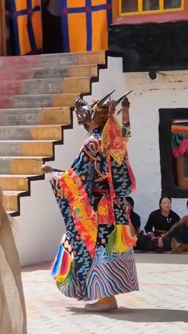 A person in a colorful, elaborate costume and mask, resembling a mythical creature with horns, dances in a courtyard. The costume features intricate patterns and vibrant colors, including blues, oranges, yellows, reds, and greens. The mask is brown and has a fierce expression with horns. The person is holding a stick in their right hand. In the background, there is a set of stairs leading up to a building with yellow and blue banners. Several people are seated on the ground in the background, observing the dance. The ground is paved with patterned tiles. The lighting suggests it is daytime, with sunlight casting shadows. A camera lens is visible in the foreground, indicating that the scene is being filmed or photographed.