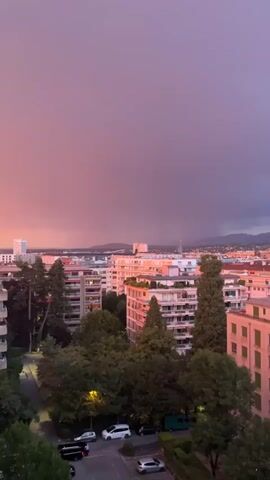 The video shows a cityscape at dusk. The sky is filled with hues of pink and purple, with a dark storm cloud on the right. In the foreground, several apartment buildings are visible, interspersed with lush green trees. A street with parked cars runs through the scene. The overall impression is one of an approaching storm over an urban area.