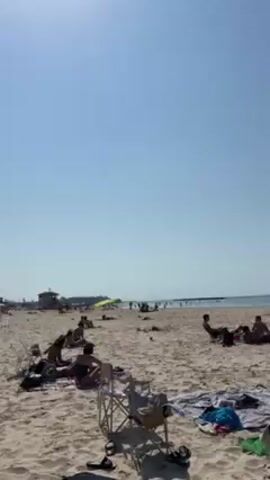 The video shows a sunny beach with many people relaxing and sunbathing. There is a tall, stone building visible on the horizon in the distance, and the sky is clear.  There are beach chairs, umbrellas, and towels scattered around. People are sitting, lying down, and walking around on the sand.