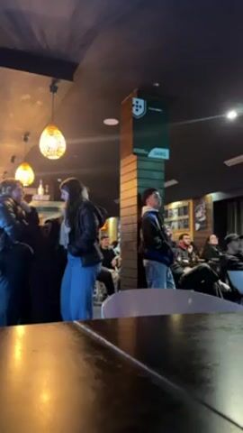 The video shows a group of people gathered in what appears to be a bar or pub, watching a game or event on a screen. Some people are standing near a bar area on the left, while others are seated in chairs facing a direction not visible in the frame. A tall, decorative pillar is in the center of the room with signage or branding on it. The interior has a modern, slightly dark ambiance with hanging lights and wall decorations. There is a Red Bull sign visible in the background. The camera is positioned low, showing a view over a dark, possibly wooden table. The overall atmosphere suggests a casual and engaged audience watching something together.
