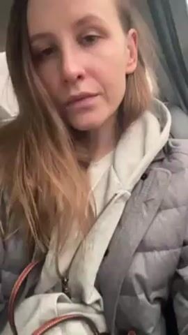 A woman with light skin and light brown hair is sitting in a car. She is wearing a white shirt, a gray hooded sweatshirt, and a gray quilted jacket. She is picking at her teeth with her fingers. The background is blurry, but it appears to be the interior of a car.