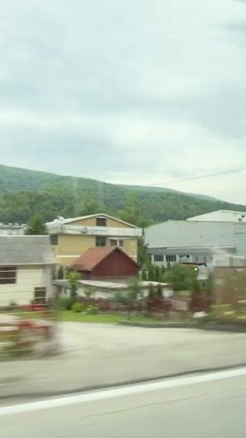 This video captures a scenic drive through Hadžići, Bosnia and Herzegovina, under a cloudy sky. The landscape is dominated by rolling green hills covered in dense forests, suggesting a rural or semi-rural setting. The road appears to be a paved highway, indicated by the white lane markings visible in the foreground.

As the vehicle moves, several buildings come into view. One prominent structure is a light yellow building with a red section, appearing to be a house or a smaller commercial building, surrounded by manicured greenery and small evergreen trees. Further along, a larger, modern industrial-style building with a light gray facade and yellow accents is visible. This building has numerous windows and appears to be a factory or warehouse. Cars are parked in front of and around this larger building, suggesting it is an active place of business.

The time of day seems to be daytime, given the ambient light, but the overcast conditions create a diffused, soft light. The weather appears to be cloudy, contributing to the muted colors in the scene. The motion blur in the video indicates that the footage was filmed from a moving vehicle, likely a car or bus, offering a transient view of the passing scenery. There are no people directly visible in the frame, and the overall mood is tranquil and observational, focusing on the natural beauty of the landscape and the presence of human structures within it. No visible text is present in the video.