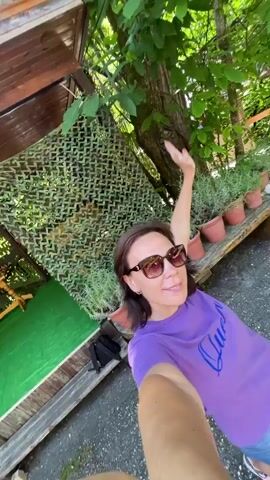 A woman with shoulder-length brown hair and sunglasses, wearing a purple t-shirt and blue jeans, takes a selfie video outdoors. She is standing in a wooded area with a wooden structure and green camouflage netting in the background. She smiles and makes a peace sign with her fingers in the first frame. The video then rotates, showing more of the wooded area, a bench with plants, and a gravel path. She continues to smile and wave.