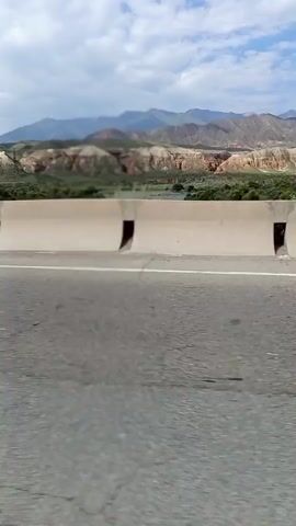 A video shows a scenic view of mountains and a river, captured from a moving vehicle. The vehicle is on a paved road separated by a concrete barrier. The landscape is arid and mountainous, with sparse vegetation. The sky is cloudy. In the background, some dust is visible, possibly from activity on a road or construction. The video pans slightly, revealing more of the mountainous terrain and the river flowing through it. A bus and a silver station wagon are briefly visible on the road.