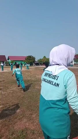 A group of students wearing green uniforms are playing on a grassy field under a clear sky. The students appear to be participating in a race or a similar athletic activity. In the foreground, a student wearing a headscarf and a green shirt with "SMP NEGERI 1 SUNGAISELAN" written on the back is facing away from the camera. In the background, there are buildings that appear to be classrooms or school facilities. A man in a black and white outfit is standing on the left side of the frame, possibly observing the students. The field is covered in dry grass, and there are shadows cast by unseen objects. The overall scene suggests an outdoor school event or physical education class.