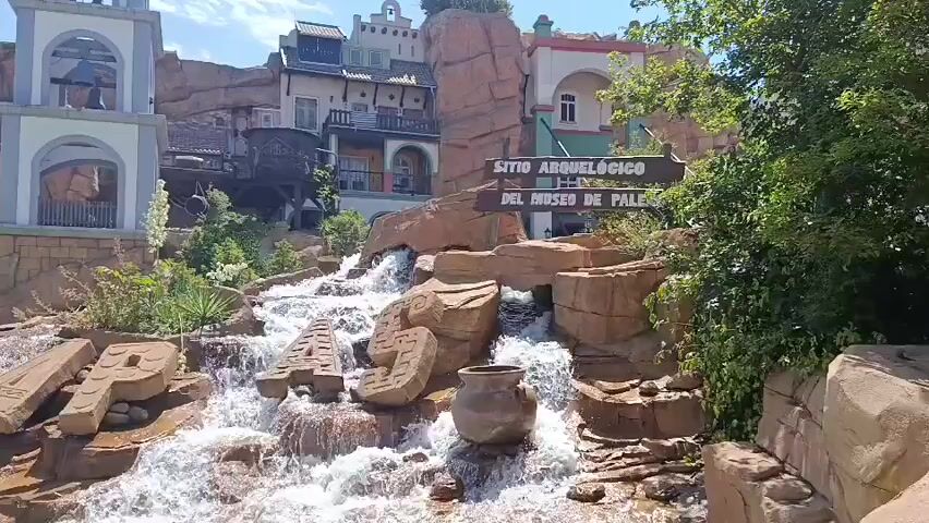 The video shows an artificial waterfall with large stone letters spelling out "APAPA" in the foreground. The letters are partially submerged in the rushing water. Behind the waterfall, there are buildings that resemble a Spanish colonial town, with a tall bell tower on the left. A wooden sign with Spanish text, "SITIO ARQUEOLOGICO DEL MUSEO DE PALENQUE," is visible on the right. The environment appears to be an outdoor theme park or attraction, with lush greenery and rocks creating a rugged, naturalistic setting. The sky is clear and blue, suggesting a sunny day.