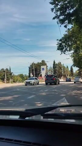 This media file captures a street view from inside a vehicle, likely during daytime. The scene appears to be Alushta, Ukraine, given the context. A prominent monument or sign with the word "АЛУШТА" (Alushta) in large red letters is visible in the background. Above the text, a blue and white emblem is displayed on a modern, abstract structure. The monument is flanked by decorative elements that resemble fans and are adorned with red, white, and blue flags, possibly the Ukrainian flag.

Several cars are on the road, including a dark colored SUV and a blue sedan that passes the monument. Trees and greenery line the street, suggesting a relatively well-maintained urban or suburban environment. Overhead, power lines and what appear to be trolleybus wires crisscross the sky, indicating public transportation infrastructure. The sky is blue with some scattered clouds, and the lighting suggests it's a sunny day. The overall mood is that of everyday movement and a glimpse of a local landmark in a coastal town, likely during pleasant weather conditions.