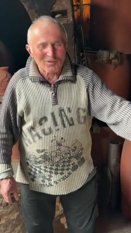 An older man is standing inside a building. He is smiling and gesturing with his hands as he speaks. He has short, light gray hair and is wearing a striped sweater with a racing-themed design. The sweater has a partial zipper on the collar. Behind him are clay pots and other pottery items. There is also a dark opening that looks like a furnace or kiln. The building appears to be a workshop or studio related to pottery. The lighting is a mix of natural and artificial. The overall impression is that he is explaining something related to his craft.