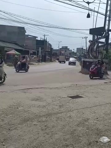 A wide shot shows a two-lane road with a dirt shoulder. On the left side of the road are several buildings, some with awnings. A red scooter with two passengers, a white scooter with two passengers, and a motorcycle with one passenger are visible on the road. A silver car is driving on the road. Overhead power lines crisscross the sky. The weather appears to be clear and sunny. The video is shot from a stationary position on the dirt shoulder.